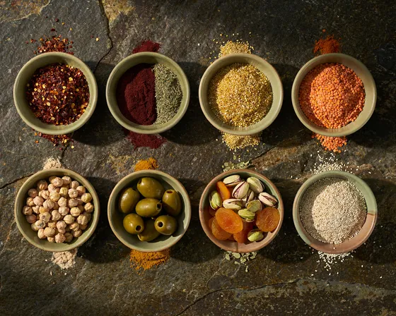 Turkish and Mediterranean pantry staples in plain ceramic bowls on stone: chilli flakes, sumac, dried oregano, bulgur wheat, red lentils, chickpeas, green olives, dried apricots, pistachios and sesame seeds.